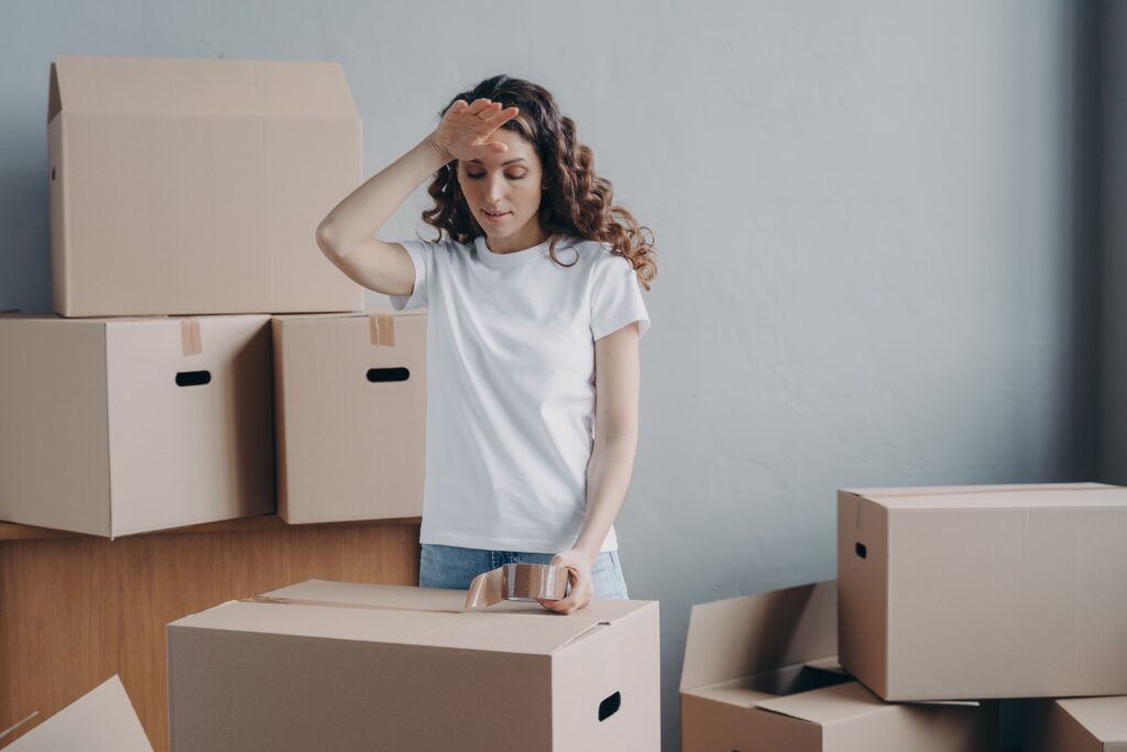 Person standing among several cardboard boxes, holding a roll of packing tape, with one hand raised to their forehead.